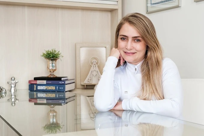Dra. Carolina de Souza Luciano em seu consultório, apoiada à mesa e sorrindo.