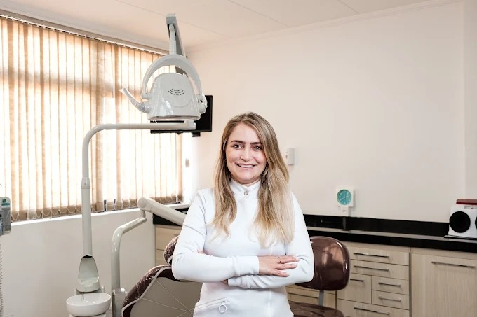 Dra. Carolina sorrindo em frente à cadeira odontológica do consultório.